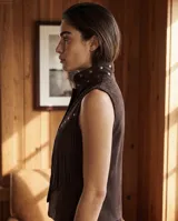 Chaleco de gamuza color marrón chocolate, sin mangas, con cuello mao, detalles de tachas metálicas en el cuello y la solapa frontal, y flecos delanteros de estilo western. Cuenta con cierre frontal de broches a presión.