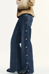 Pantalón de jean azul oscuro, de corte amplio y tiro medio, con bolsillos delanteros y traseros. Presenta una hilera de botones metálicos a lo largo de cada lateral.