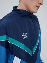 Campera deportiva Umbro modelo Beat, confeccionada en nylon ligero con diseño de bloques de color en blanco, azul marino y verde azulado. Presenta cierre frontal, cuello alto y elásticos en puños y cintura.