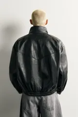 Cazadora relaxed fit de tejido efecto piel en color negro, con cuello subido, manga larga, bolsillos con solapa en el delantero y acabados elásticos. Cierre frontal con cremallera oculta por solapa.