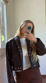 Campera corta de ecocuero color marrón chocolate, con diseño de solapas decoradas con tachas metálicas plateadas y cierre frontal.