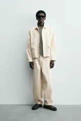 Cazadora de hombre color beige claro, corte relajado, con cuello solapa, manga larga y puños con botones a presión. Presenta bolsillos de plastrón con solapa en la cadera y cierre frontal de cremallera oculta por solapa de botones a presión.