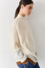 Buzo tejido de punto con cuello alto, diseño holgado y aberturas laterales. Presenta un cuerpo en color azul melange con detalles de franjas en color crema y verde en los puños y el ruedo. Incluye un pequeño bordado en el pecho.