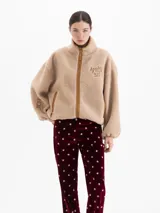 Campera de tejido polar color beige con cuello alto, cierre frontal y bolsillos laterales con cierre. Presenta bordado 'APRÈS SKI' en el pecho y detalles en color tostado en los bordes. Diseño de corte holgado con mangas largas.