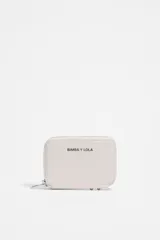 Monedero rectangular de cuero color blanco, con cierre de cremallera perimetral y tirador metálico. Presenta el logo de la marca Bimba y Lola estampado en el frente y detalles de ribetes metálicos en la base. Interior con compartimentos para tarjetas.
