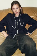 Blusa de manga larga en color azul marino, con cuello solapa y lazo frontal. Presenta detalles de pespuntes bordados en color crema a lo largo del cuello, lazo, cierre frontal y puños. Cierre frontal con botones.