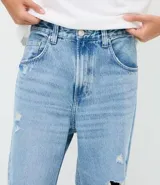 Pantalón de jean azul claro con corte baggy relaxed, cintura con pasantes, cierre frontal con botón y roturas localizadas en las piernas.
