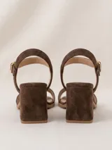 Sandalias de gamuza color marrón chocolate, de taco cuadrado medio y punta cuadrada. Tienen una tira delantera ancha y una tira ajustable al tobillo con hebilla metálica dorada.