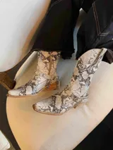 Botas de caña media con estampado de piel de serpiente en tonos beige y marrón. Diseño estilo cowboy con punta cuadrada, detalle de hebilla y apliques metálicos.
