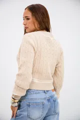 Sweater de punto trenzado en color beige, con cuello en V y detalles de rayas blancas en el cuello, puños y ruedo. De corte ajustado y manga larga.