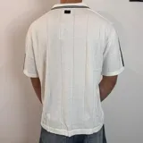 Chomba de tejido de punto color negro, con cuello polo y cierre frontal parcial. Presenta diseño de rayas verticales sutiles en el tejido, detalles de líneas blancas en los hombros y estampado frontal con texto y gráficos en color blanco.