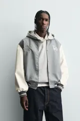 Cazadora estilo varsity o bomber, cuerpo principal gris oscuro y mangas color crema. Presenta cuello, puños y bajo en rib elástico con rayas grises y blancas. Tiene bordados en el pecho: un monograma estilizado a la izquierda y el texto "EST. 1975" a la derecha.