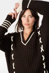 Sweater de punto estilo preppy en color crudo con detalles en marrón. Presenta cuello en V con ribete a rayas, tejido con textura de ochos y cordones laterales decorativos. Posee puños y ruedo acanalados con diseño a rayas.