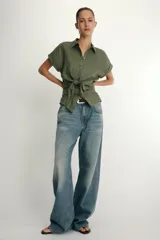 Camisa blanca confeccionada en tejido con lino, con cuello solapa y manga muy corta con terminación en vuelta. Presenta un detalle de nudo en el delantero y cierre frontal con botones metálicos.