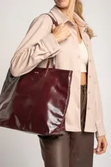 Bolso tipo tote de gran tamaño en color bordó con acabado brillante. Cuenta con doble asa de hombro y placa metálica decorativa con logo en la parte frontal.