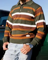 Buzo tejido de punto con diseño de rayas horizontales en tonos verde, marrón y blanco. Presenta cuello tipo polo con cierre de tres botones, mangas largas con puños acanalados y logo de la marca bordado en el pecho. Incluye una etiqueta decorativa en la parte inferior.