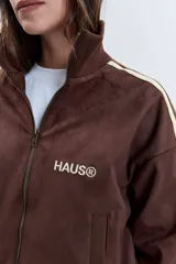 Cazadora de estilo deportivo en color marrón, confeccionada en tejido tipo gamuza. Presenta cierre frontal completo, cuello alto, puños y ruedo acanalados, y franjas laterales en tono rosado. Incluye el logo de la marca bordado en el pecho.