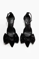 Zapatos de tacón tipo pump en terciopelo negro, con punta fina y un lazo grande decorativo en el empeine. Presentan una pulsera ajustable al tobillo con cierre de hebilla y tacón fino forrado.