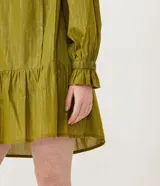 Vestido corto de manga larga en color verde oliva, confeccionado en tejido plano con hilos metalizados a contraste. Presenta cuello redondo con lazo decorativo y flecos, mangas largas con puños elásticos y detalle de volados en los puños.