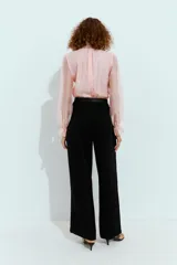 Mono largo con parte superior de blusa rosa de manga larga, cuello alto fruncido y puños con volantes. La parte inferior es un pantalón negro de pernera ancha con bolsillos delanteros y un cinturón negro con hebilla dorada.