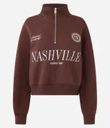 Buzo de algodón y poliéster color marrón, con cuello alto y cierre frontal parcial. Presenta diseño de hombros caídos, puños y ruedo acanalados, y estampado tipográfico frontal con la palabra Nashville.