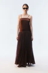 Vestido largo color marrón, con breteles finos y escote recto. La parte superior es lisa, con una falda larga y amplia de pliegues finos que aporta movimiento.