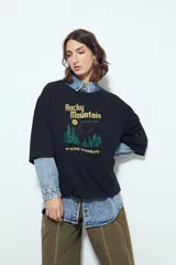 Remera blanca de manga corta con calce relajado y cuello a la base. Presenta un estampado frontal pequeño y una estampa protagonista en la espalda con el texto "Rocky Mountain National Park" y un paisaje de montañas y árboles.