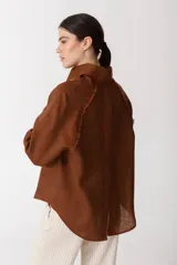 Camisa color ladrillo con rayas verticales finas color crema, de corte recto y mangas largas con puños abotonados. Presenta cuello camisero, cierre frontal con botones y detalle de flecos en los hombros.