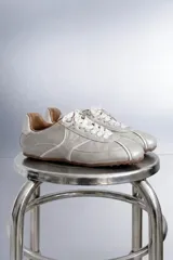 Zapatilla baja de microfibra color gris plateado, con diseño aerodinámico, costuras pespuntadas en contraste blanco, cordones blancos y suela de goma marrón con textura de tapones.
