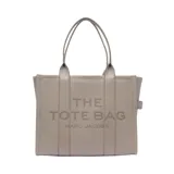 Bolso tipo tote de gran tamaño, confeccionado en cuero marrón con textura granulada. Presenta el logo de la marca estampado en el frente con la inscripción The Tote Bag Marc Jacobs. Cuenta con asas superiores fijas y un diseño estructurado y espacioso.