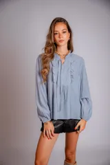 Camisa de color celeste con volados decorativos en el cuello y la parte delantera, cuenta con cordones para ajustar el escote, mangas largas con puños y corte holgado.