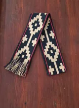 Faja tejida en telar de mano con diseño geométrico en tonos negro, beige y rojo, con terminación de flecos en los extremos.