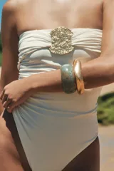 Bañador enterizo tipo bandeau color crema, con escote recto drapeado y detalle de pieza metálica circular dorada en el centro del pecho. Cuenta con forro interior.