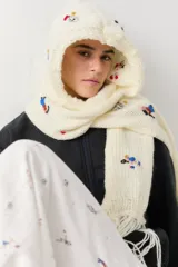 Bufanda con capucha integrada, confeccionada en tejido de punto grueso color crudo. Presenta bordados coloridos de figuras deportivas y cuenta con flecos en los extremos.