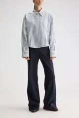 Camisa de calce relajado confeccionada en algodón, con diseño de rayas verticales en tonos beige y blanco. Presenta cuello clásico, cierre frontal con botones, puños abotonados y largo ligeramente cropped.