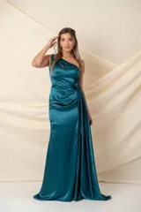 Vestido largo de satén color verde petróleo, con diseño asimétrico de un solo hombro y abertura frontal en el escote. Presenta una cintura drapeada con detalle de flor lateral, falda con tajo frontal y cierre de botones forrados en la espalda.