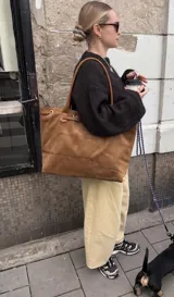 Cartera tipo tote de símil gamuza color marrón, con diseño estructurado y amplio, asas de hombro delgadas y detalle de tira con hebilla y argollas metálicas en la parte superior.