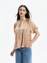 Blusa de satén color tostado con cuello redondo, cierre frontal con cordones ajustables y mangas cortas tipo raglán con diseño holgado.