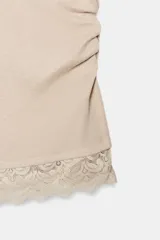 Camiseta ajustada (slim fit) color crema, con cuello redondo y mangas sisa. Presenta frunces laterales y un bajo asimétrico con detalle de encaje a tono.