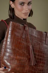 Bolso tipo tote de cuero vacuno con textura de reptil, color marrón. Presenta detalles de pasamanería en los bordes laterales y una borla de flecos de cuero colgante. Cuenta con cierre superior.