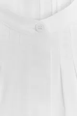 Camisa blanca de algodón con cuello redondo y manga corta abullonada. Presenta un canesú con pliegues verticales en la parte delantera y cierre frontal con botones ocultos. El bajo es redondeado.