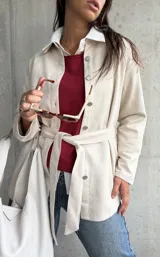Sobrecamisa de gamuza en color beige, con cierre frontal de botones metálicos, cuello camisero y lazo ajustable en la cintura. Presenta un diseño de corte recto y mangas largas.