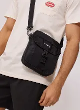 Bandolera Rusty modelo Vansi de color negro, con diseño estructurado, bolsillo frontal con solapa y cierre de hebilla plástica, correa regulable y removible, y etiqueta con el logo de la marca en el frente.