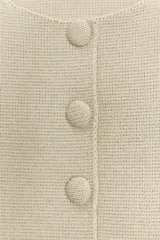 Chaqueta de punto color beige claro, con cuello redondo, manga corta y bajo peplum con pliegues. Cierre frontal con botones forrados a tono.