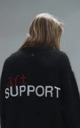 Sweater de punto negro con textura suave, cuello redondo y diseño oversize. Presenta un bordado frontal con la frase 'art SUPPORT' en letras rojas y blancas, y un pequeño detalle de tijeras bordado en la nuca.