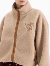 Campera de tejido polar color beige con cuello alto, cierre frontal y bolsillos laterales con cierre. Presenta bordado 'APRÈS SKI' en el pecho y detalles en color tostado en los bordes. Diseño de corte holgado con mangas largas.
