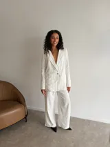 Pantalón de corte sastrero y pierna ancha, color beige con diseño de rayas verticales finas. Presenta pinzas en la cintura, cierre frontal con botón y bolsillos laterales.