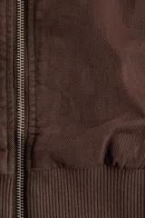 Campera bomber color marrón oscuro de tejido grueso, con cuello corto, puños y bajo acanalados. Cuenta con cierre frontal plateado, bolsillos frontales insertados y diseño de hombros caídos. Está completamente forrada.