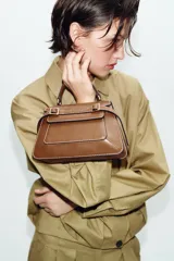 Bolso de mano tipo city color marrón, con diseño estructurado, costuras a la vista en color blanco y detalles de hebillas metálicas decorativas en la parte frontal. Cuenta con asa de mano superior, correa ajustable para usar como bandolera y cierre mediante solapa con imán.