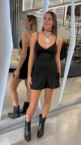 Vestido corto negro de tirantes finos, con escote en V y detalle de frunces en la cintura. La falda presenta un diseño con volado en el ruedo.